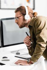Fototapeta premium Side view of handsome digital designer in headphones using computer and holding stylus of graphics tablet at table in office
