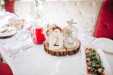Festive stands in the wedding banquet area served with dishes and cutlery and decorated with compositions of flowers, greenery, cages with candles inside and table numbers