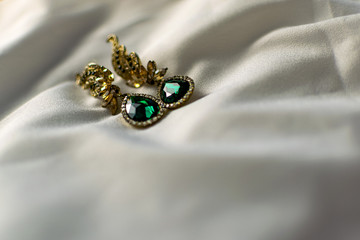 Metal gold earrings with green, emerald stones, white and yellow rhinestones. On a white fabric flattened background. Brilliant and festive decoration