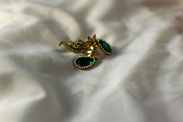 Metal gold earrings with green, emerald stones, white and yellow rhinestones. On a white fabric flattened background. Brilliant and festive decoration