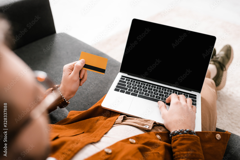 Poster selective focus of digital designer using laptop with blank screen and holding credit card on couch