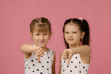 two cute little child girl in dress showing the thumbs down gesture on pink background.