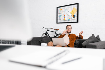 Selective focus of smiling 3d artist talking on smartphone and holding laptop on couch in office