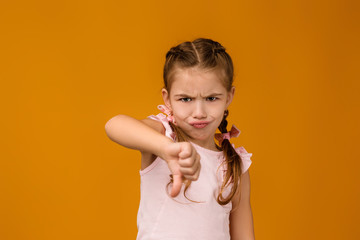 Cute little child girl in dress showing the thumbs down gesture on yellow background.