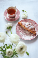 A morning table with a cup of tea with croissant and white ranunculus flowers blurred 