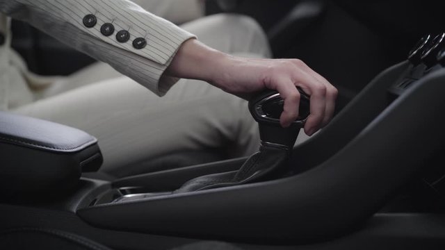 Close-up of female Caucasian hand holding gear shift. Unrecognizable young businesswoman in elegant white suit sitting on driver's seat in vehicle. Auto industry, lifestyle, wealth, car dealership.