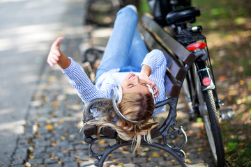 Young blonde woman in the park lying on bench and listen music over headphones with bycicle