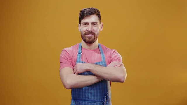 Bearded attractive caucasian man chef wearing blue striped apron shakes off flour powder cleaning after crazy baking in the kitchen posing happy on orange background.