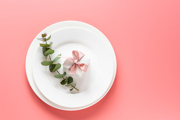 Beautiful festive composition with white plates, gift box with bow and eucalyptus twig on pink pastel background. Invitation card concept. Table setting. 
