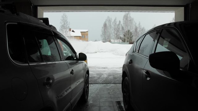 Man cleans snow with a snow thrower. View from two car from garage