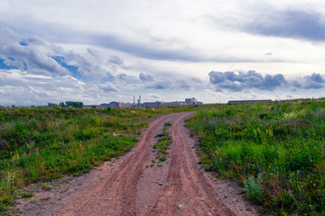 Summer landscape dirt road on a background of cloudy sky and urban buildings on the horizon.