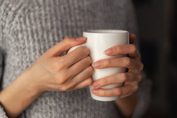 A white mug in the hands of a girl in a gray sweater. He warms his hands.