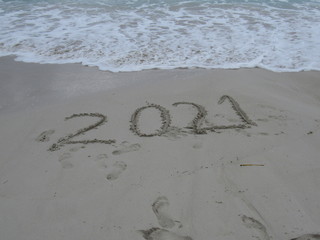 Happy New Year 2021, lettering on the beach with wave and clear blue sea. Numbers 2021 year on the sea shore, message handwritten in the golden sand on beautiful beach background. New Years concept.