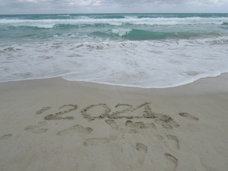 Happy New Year 2021, lettering on the beach with wave and clear blue sea. Numbers 2021 year on the sea shore, message handwritten in the golden sand on beautiful beach background. New Years concept.