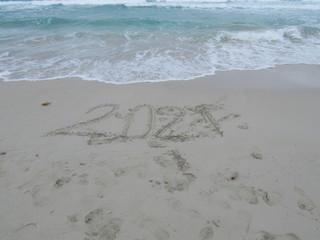 Happy New Year 2021, lettering on the beach with wave and clear blue sea. Numbers 2021 year on the sea shore, message handwritten in the golden sand on beautiful beach background. New Years concept.