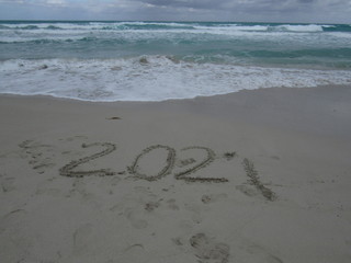 Happy New Year 2021, lettering on the beach with wave and clear blue sea. Numbers 2021 year on the sea shore, message handwritten in the golden sand on beautiful beach background. New Years concept.