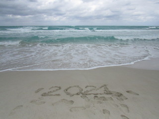Happy New Year 2021, lettering on the beach with wave and clear blue sea. Numbers 2021 year on the sea shore, message handwritten in the golden sand on beautiful beach background. New Years concept.