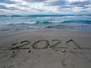 Happy New Year 2021, lettering on the beach with wave and clear blue sea. Numbers 2021 year on the sea shore, message handwritten in the golden sand on beautiful beach background. New Years concept.
