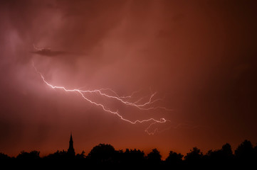 Rain falling and stroke of forked lightning during thunderstorm at night over church tower and trees