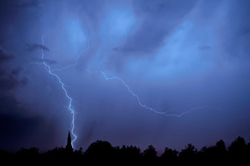Rain falling and stroke of forked lightning during thunderstorm at night over church tower and trees