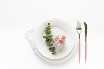 Festive table setting for romantic dinner with gift box on a plate, cutlery an eucalyptus twig on white background. Invitation card concept.