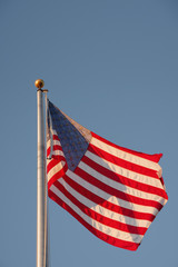 A US American flag waving high up on a metal flagpole in the sunshine under bright blue sky