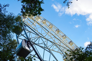 Bottom view of ferris wheel through green tree on background of blue sky in sunny summer day.