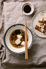 Japanese Miso broth soup with silk tofu cubes, soy beans edamame, green beans in traditional bowl with chopsticks on grey linen table cloth. Asian dinner. Flat lay, space