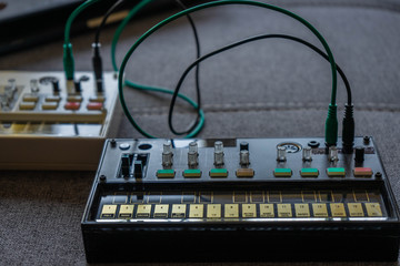 Close up view of a small black fm synthesizer with patch cables with another synth behind it on top of a grey fabric sofa. Electronic music concept