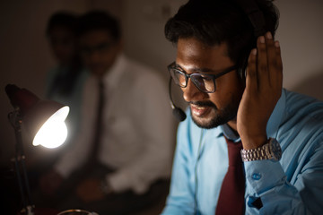 An Indian Bengali tall, dark, handsome brunette young man/tele caller in office wear is sitting on a office table in a working mood in a corporate office/bpo/call center. Indian corporate lifestyle