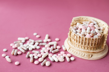 Stone statuette of the Coliseum building in Rome in Italy, surrounded by a bunch of antiviral pills and tables. Concept of dangerous infection with the Chinese coronavirus covid-2019. Pink background