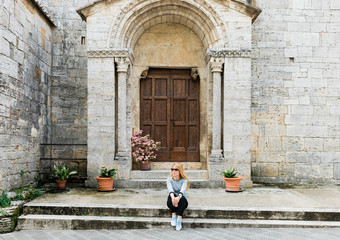 Young female traveler sitting on steps of medieval church in Tuscany, Italy 