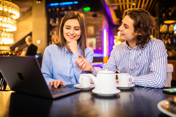 Beautiful young couple is using a laptop, talking and smiling while sitting in the cafe.