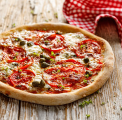 Homemade pizza with addition tomatoes  cheese and capers on a rustic wooden board, close up