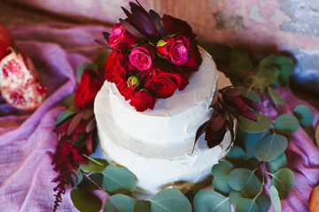 White wedding cake with red flowers and eucalyptus leaves on a table covered with pink fabric.
