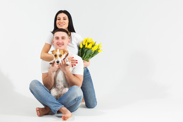 Close up portrait young couple sit and hugging, holding yellow flowers and dog in studio on white background. couple embracing with dreamy amorous expression. Lovely family. Celebrating woman's day.