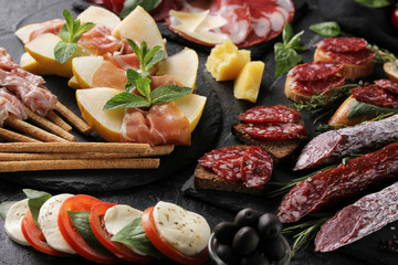 The concept of Italian cuisine. Meat appetizers with herbs, caprese and melon. Pancetta, coppa, prosciutto and italian sausage on a black background
