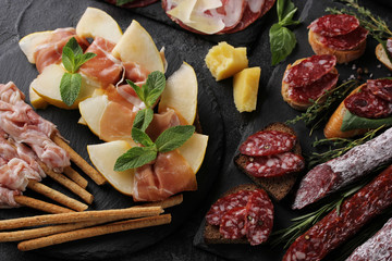 The concept of Italian cuisine. Meat appetizers with herbs and melon. Pancetta, coppa, prosciutto and italian sausage on a black background