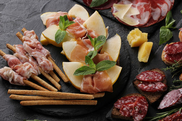 The concept of Italian cuisine. Meat appetizers with herbs and melon. Pancetta, coppa, prosciutto and italian sausage on a black background