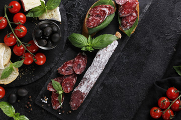 The concept of Italian cuisine. Sausage on a black board with tomatoes, olives, basil and cheese. Bread with sausage, sandwich. Background image, copy space