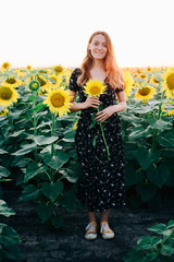 a young beautiful girl in a black floral dress stands in a field among sunflower flowers at sunset