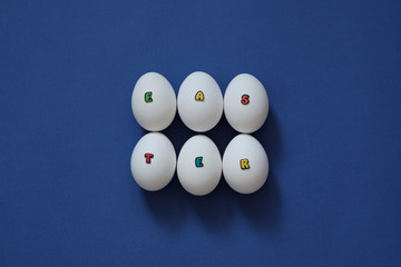 Creative layout of white chicken eggs with the inscription Easter from colored letters on a blue background. Top view, easter holidays concept
