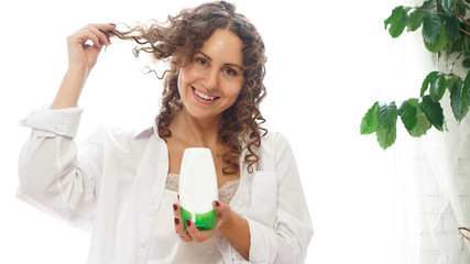 Pretty woman showing white cosmetic tube for curly hair care in her hand at home, white room with green plants