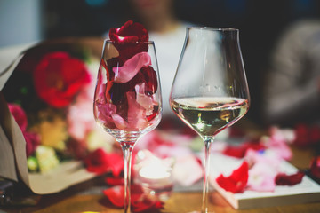 Beautiful glass with rose leafs, rose-petal, rose petals on a romantic date with wine, candles in candlelight and with celebrating couple on the background, interior for a romantic date diner