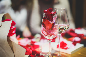 Beautiful glass with rose leafs, rose-petal, rose petals on a romantic date with wine, candles in candlelight and with celebrating couple on the background, interior for a romantic date diner