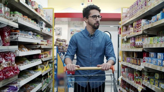 Serious man buying products in store. Focused bearded young man with shopping trolley choosing products in supermarket, tracking shot. Shopping concept