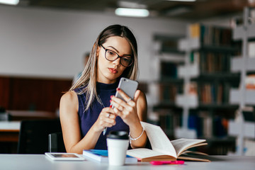 Young woman studying at table in library. Using smartphone