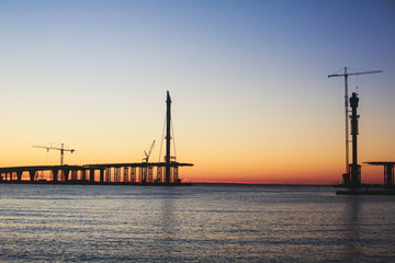 Obraz premium An unfinished part of highway road or bridge, on sunset or dusk, Industrial construction cranes and building silhouettes at sunrise, back light construction site silhouetted at sunset, over the water