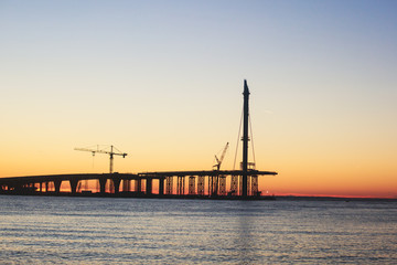 An unfinished part of highway road or bridge, on sunset or dusk, Industrial construction cranes and building silhouettes at sunrise, back light construction site silhouetted at sunset, over the water