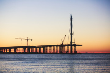 Fototapeta na wymiar An unfinished part of highway road or bridge, on sunset or dusk, Industrial construction cranes and building silhouettes at sunrise, back light construction site silhouetted at sunset, over the water